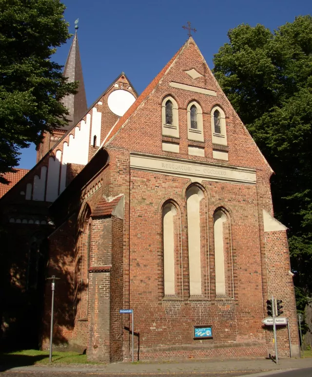 Backsteinfassade der Wittenburger Bartholomäuskirche mit hohen Fenstern; links im Hintergrund die Turmspitze zwischen Bäumen.